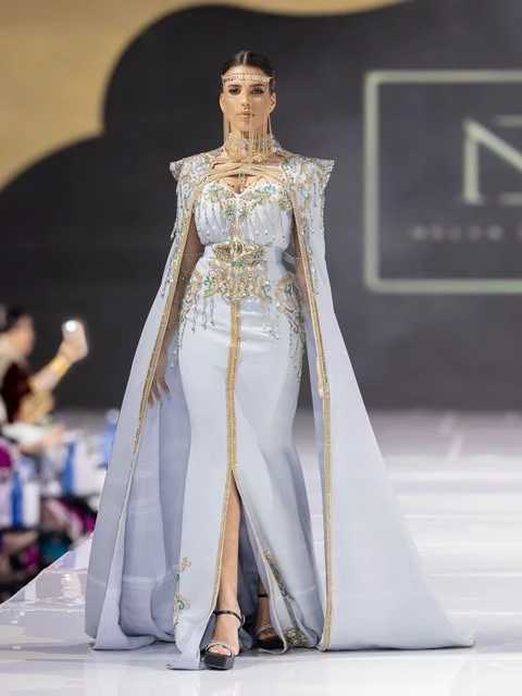 Modelo de vestido azul-gelo bordado em dourado, com capa longa e véu de correntes, na passarela